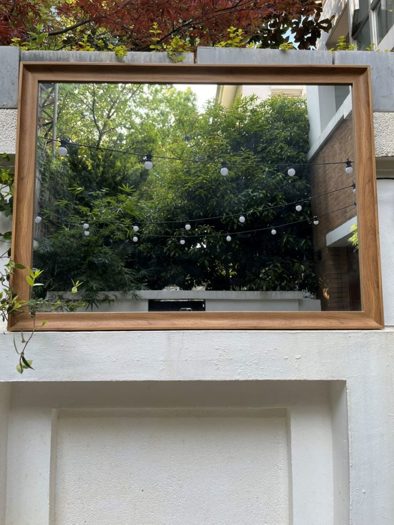 Grand miroir extérieur encadré de bois reflétant la végétation et l’architecture, créant une illusion de profondeur au jardin