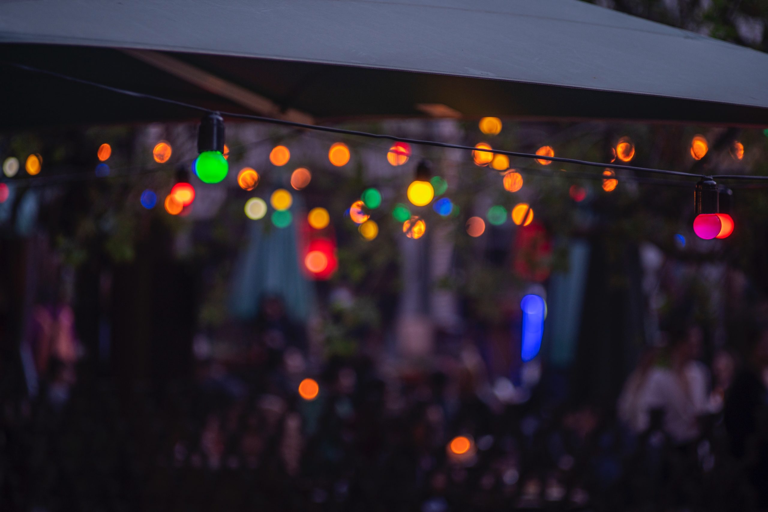 Guirlande de lumières colorées suspendue sous un auvent au-dessus d’une terrasse animée en soirée, créant une ambiance chaleureuse et conviviale.